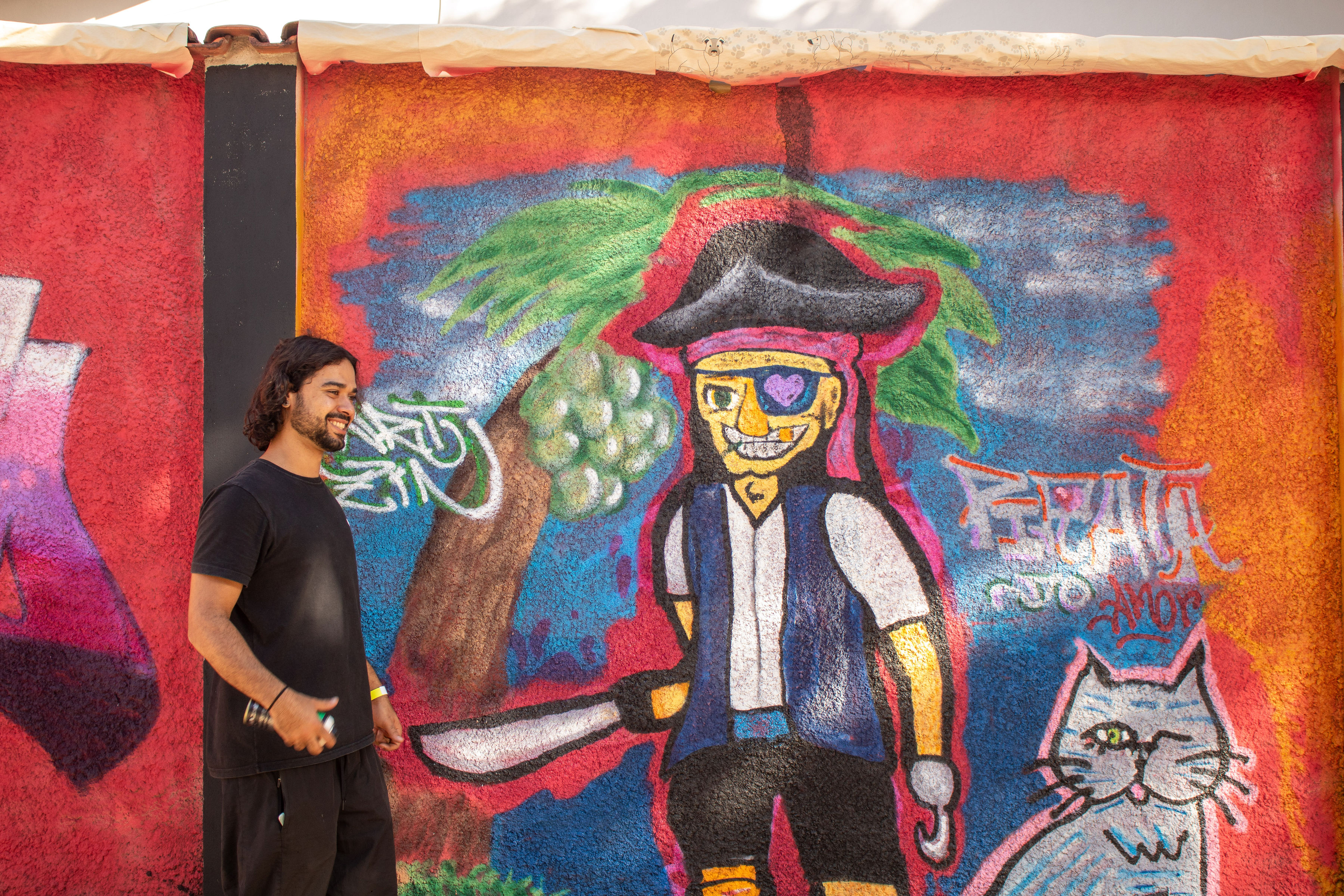 Homem de cabelos escuros e barba, vestindo camiseta e calça pretas, sorri enquanto segura uma lata de spray diante de um mural colorido com grafite de um pirata sorridente, usando tapa-olho, faixa rosa e chapéu preto, com um facão na mão e um gato ao lado. Ao fundo, há uma palmeira e um céu estilizado com cores vibrantes como vermelho, azul e laranja.