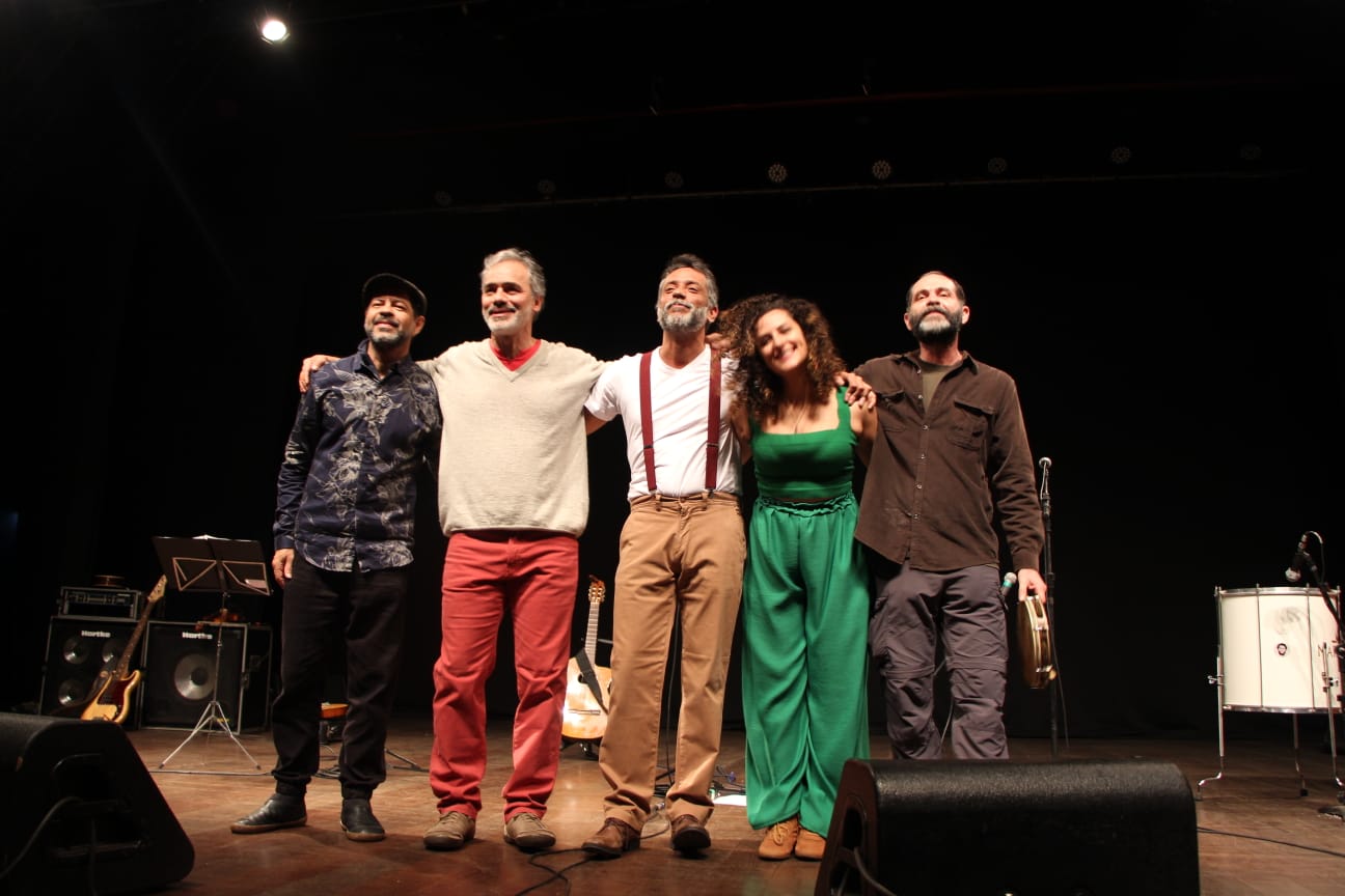 Cinco músicos estão lado a lado no palco, de braços dados, sorrindo e olhando para a plateia, como em agradecimento após uma apresentação. Ao fundo, há instrumentos musicais como violões, amplificadores e um tambor. O palco é escuro com iluminação voltada para o grupo, que está vestido com roupas variadas e descontraídas.