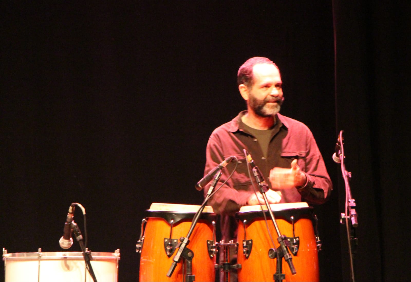 Homem de barba grisalha toca dois tambores conga em um palco escuro, iluminado por luzes quentes. Ele está sorrindo levemente, veste camisa escura e há vários microfones posicionados ao redor dos instrumentos de percussão. Ao lado esquerdo, também é visível parte de um tambor branco com microfone direcionado. A cena sugere uma apresentação musical ao vivo.