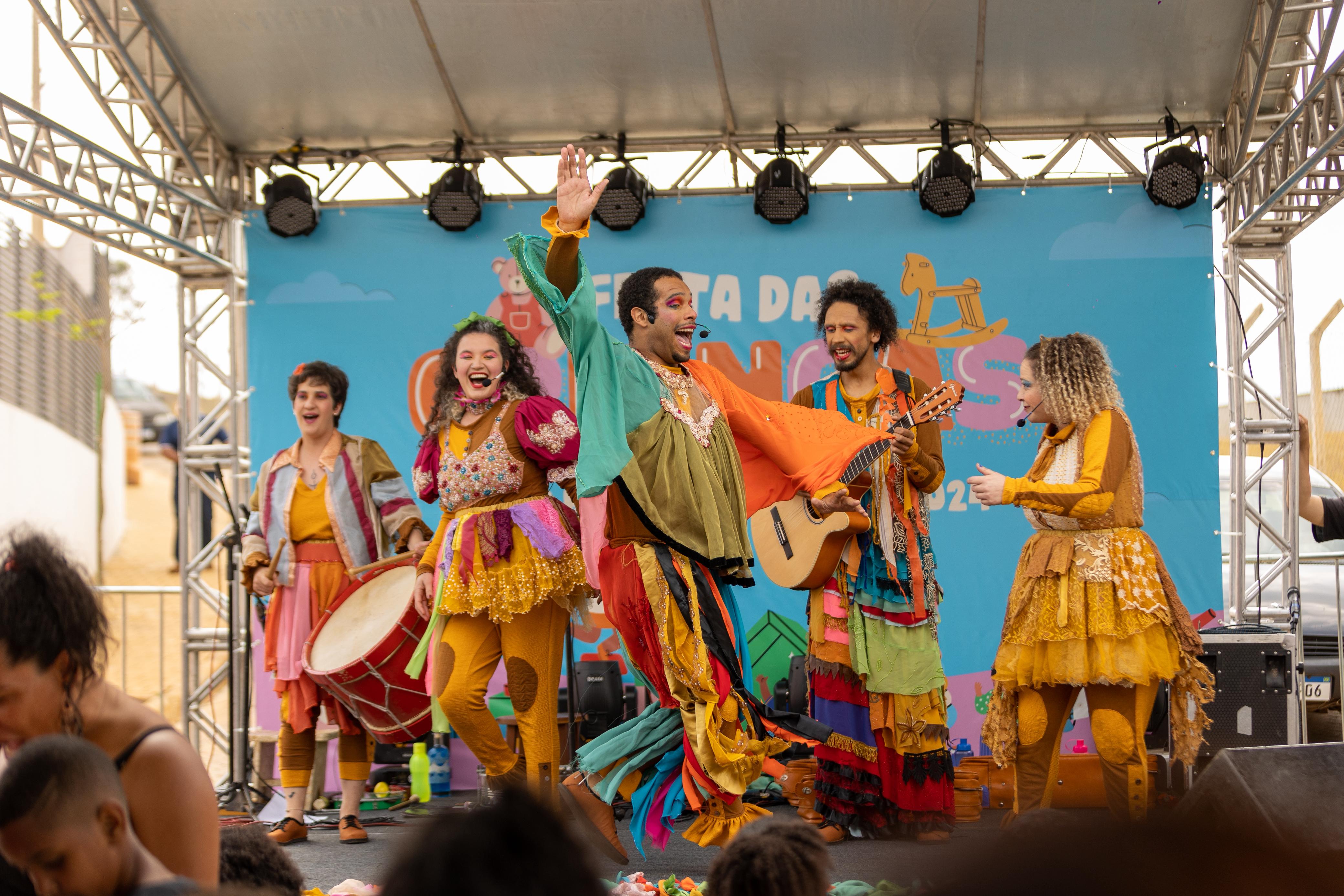 Grupo de artistas fantasiados se apresenta em um palco colorido durante um evento infantil. Eles estão cantando e dançando com alegria, usando roupas vibrantes e teatrais. Ao fundo, uma faixa azul com o texto 'Festa das Crianças'. Um dos artistas toca violão, enquanto outro levanta o braço sorrindo. Crianças assistem à apresentação em frente ao palco.