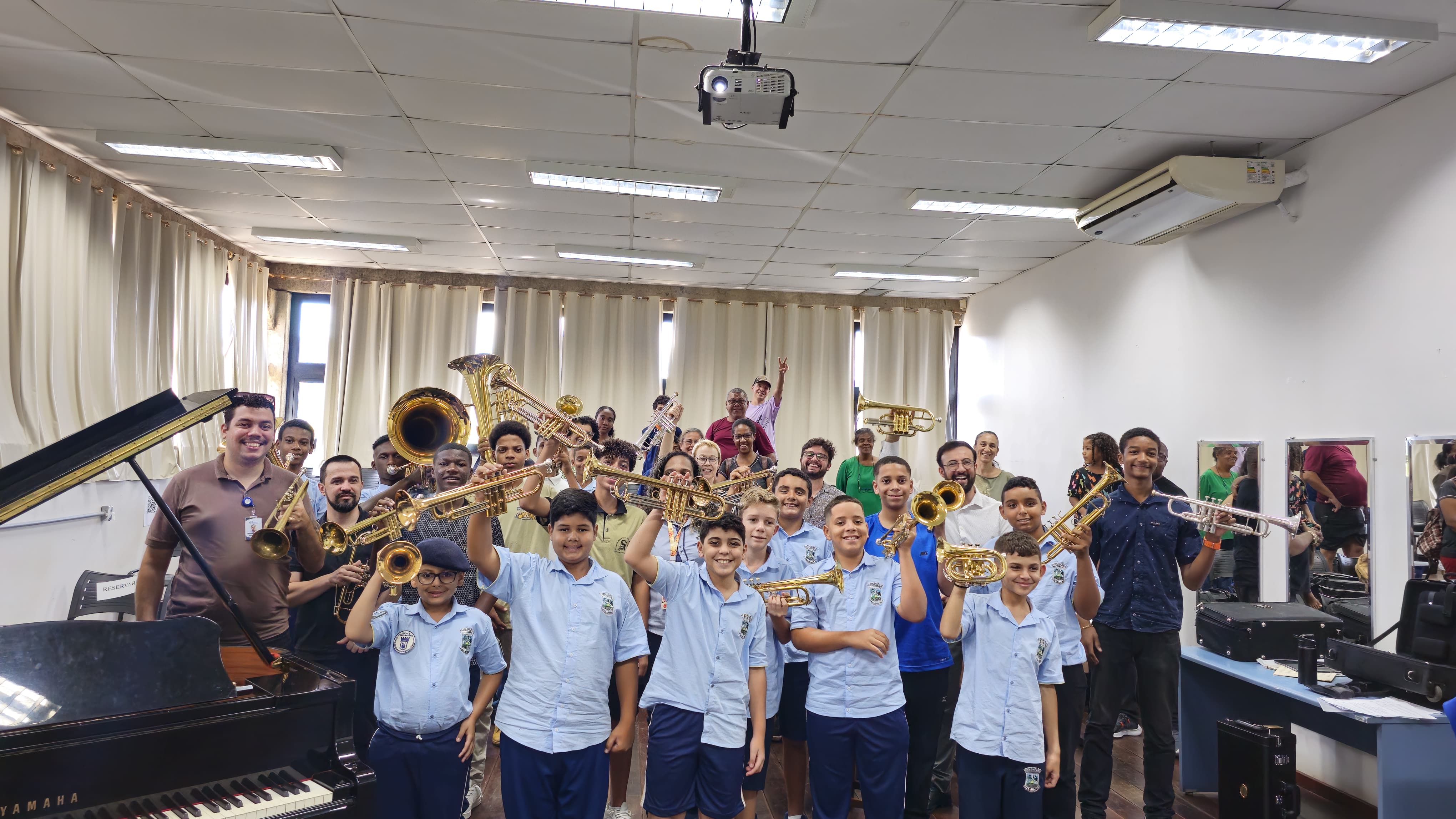 Foto de grupo em uma sala de música com dezenas de crianças e adultos sorrindo e segurando instrumentos de sopro, como trompetes, trombones e tubas. Os alunos na frente usam uniformes escolares azul claro, enquanto os adultos e outros participantes estão ao fundo, próximos a cortinas claras. Um piano de cauda aparece à esquerda e há espelhos e equipamentos musicais à direita.