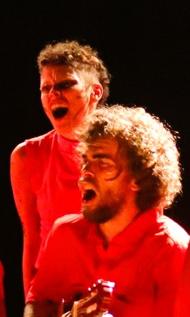 Duas pessoas em cena teatral, ambas com expressões intensas e bocas abertas como se estivessem gritando ou cantando. A pessoa ao fundo tem cabelo curto com as laterais raspadas e usa uma blusa vermelha. A pessoa em primeiro plano tem cabelo cacheado e barba, também veste vermelho e segura um instrumento musical, possivelmente um cavaquinho ou violão. A iluminação é quente, destacando tons de vermelho e laranja, e o fundo é escuro.