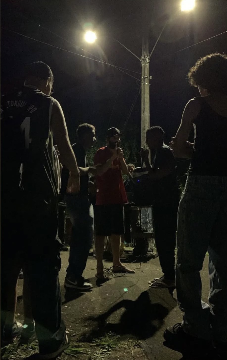 m grupo de pessoas está reunido à noite em uma rua mal iluminada, aparentemente participando de um sarau ou roda de rima. Elas estão em pé, formando um círculo sob dois postes de luz. Ao centro, uma pessoa com camiseta vermelha segura um microfone, indicando que está se apresentando. As silhuetas são realçadas pela iluminação vinda de cima, e o ambiente é urbano e informal.