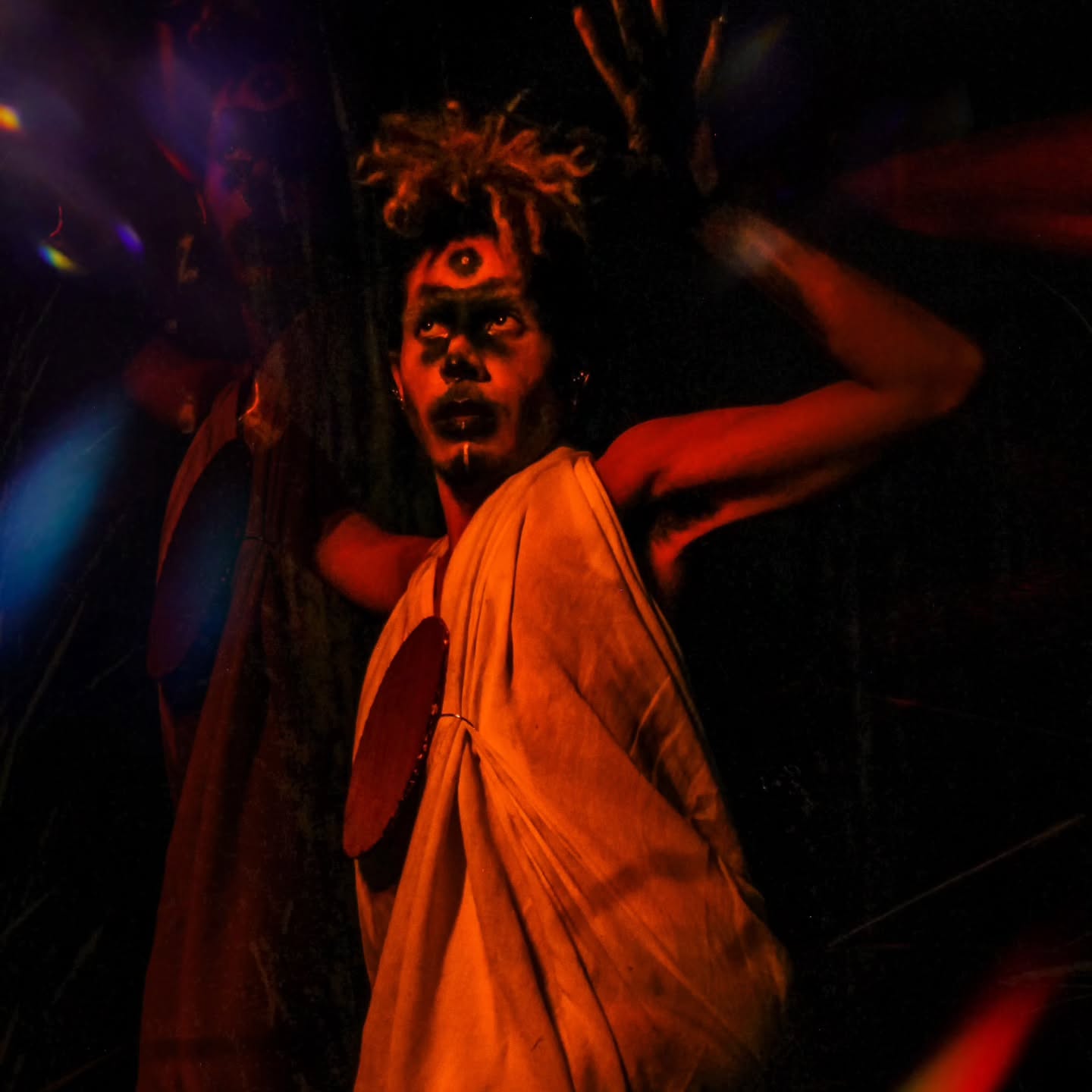 Pessoa em performance teatral ou ritualística, iluminada por luz avermelhada e envolta em sombras. Veste túnica clara com dois círculos escuros aplicados e tem maquiagem marcante, com destaque para um terceiro olho pintado na testa. A expressão é intensa, com os olhos voltados para cima e os braços erguidos. Reflexos e feixes de luz coloridos atravessam a cena, criando um efeito onírico e misterioso. Ao fundo, uma figura espelhada ou duplicada adiciona à atmosfera surreal da imagem.