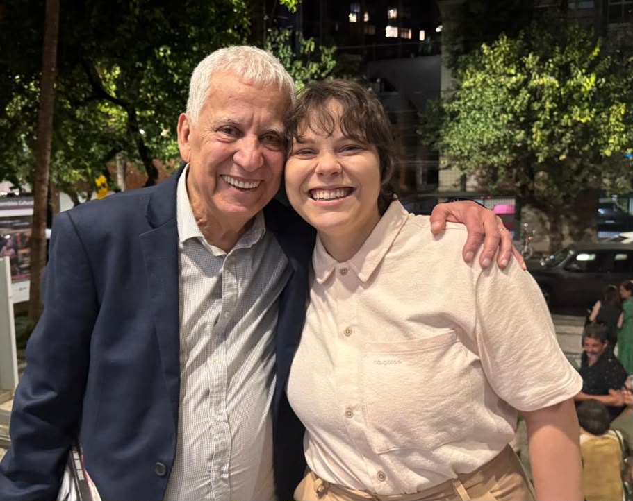 Fotografia noturna ao ar livre de duas pessoas abraçadas e sorrindo para a câmera. À esquerda, um homem idoso de cabelos brancos, vestindo paletó azul e camisa clara, está com o braço no ombro da pessoa ao lado. À direita, uma jovem de cabelos curtos escuros veste uma camisa clara de botões. Ao fundo, um pouco desfocados, há árvores, rua, carros e pessoas em uma calçada.