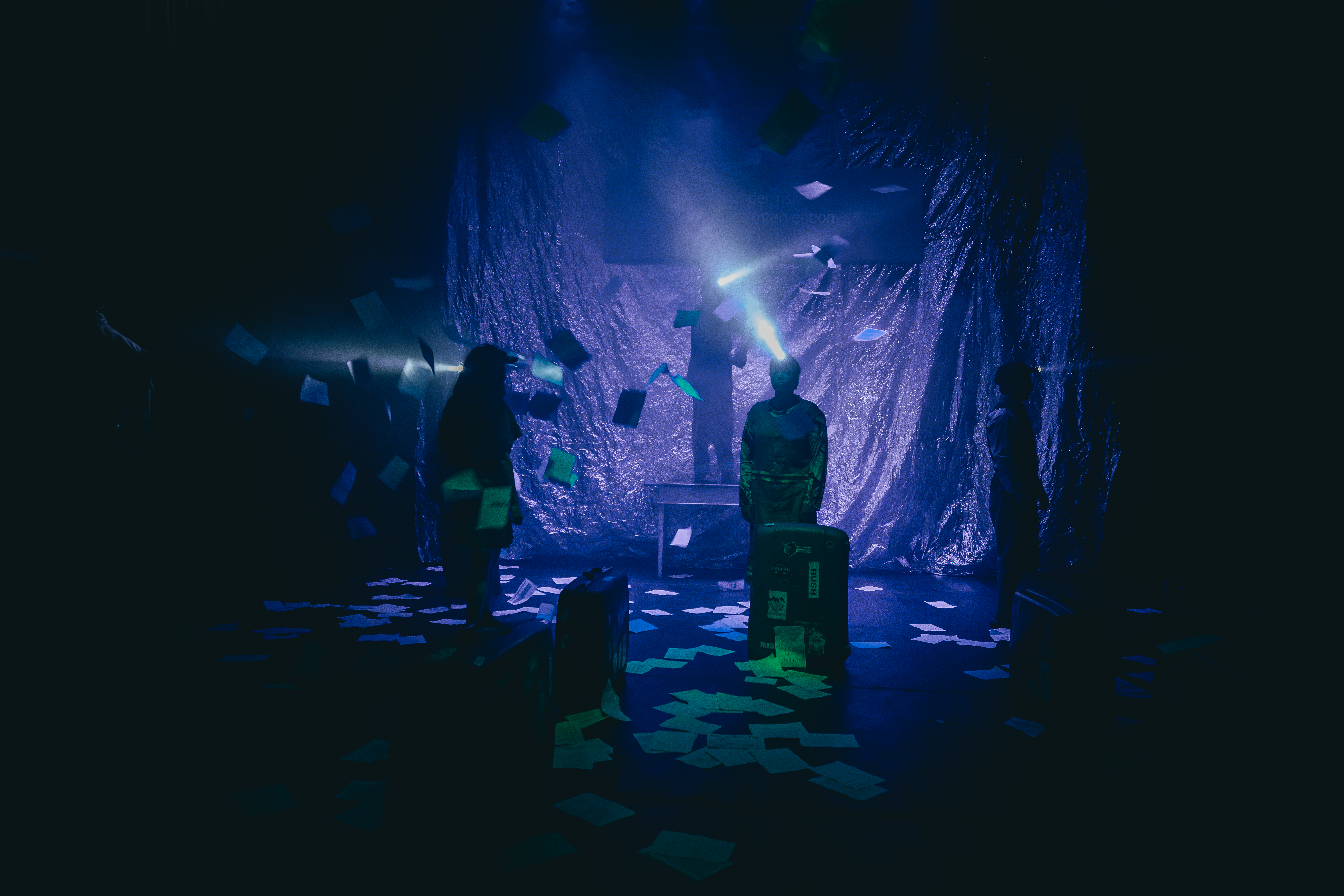 Cena de performance teatral em um palco escuro com luz azul. O fundo é uma cortina reflexiva. Inúmeros papéis estão espalhados pelo chão e suspensos no ar, com brilhos esverdeados. Três performers são vistos como silhuetas. Duas delas têm lanternas de cabeça projetando feixes de luz. A figura central está em pé sobre malas e uma mesa. Malas de viagem estão pelo palco. Atmosfera dramática e misteriosa.