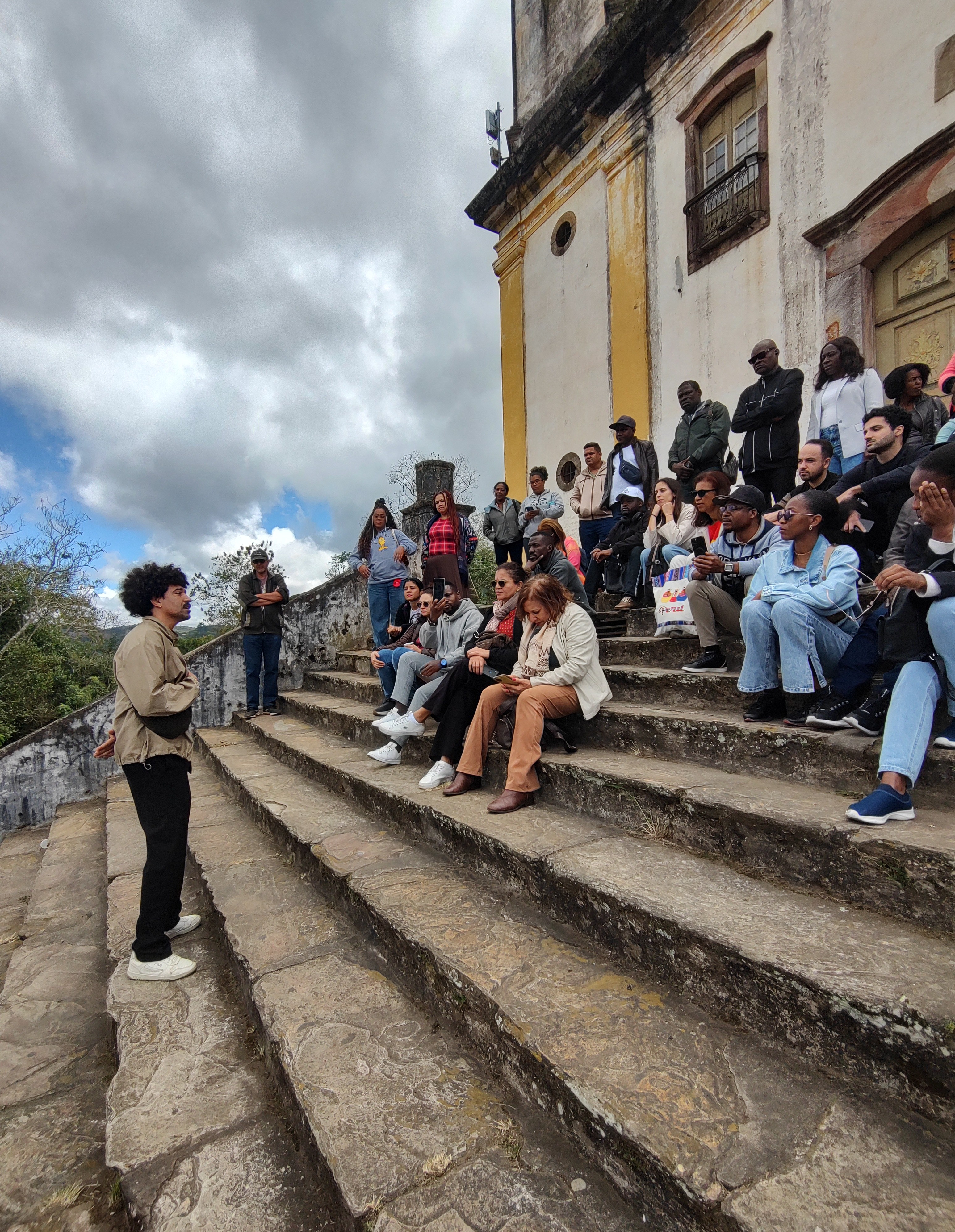 Fotografia ao ar livre. À esquerda, um homem negro com cabelo crespo, vestindo jaqueta bege e calça preta, está em pé nos degraus de pedra, falando para um grupo. À direita, dezenas de pessoas estão sentadas e em pé na ampla escadaria de pedra de uma igreja colonial antiga, de paredes brancas e amarelas, prestando atenção nele. O céu ao fundo está nublado com aberturas azuis.