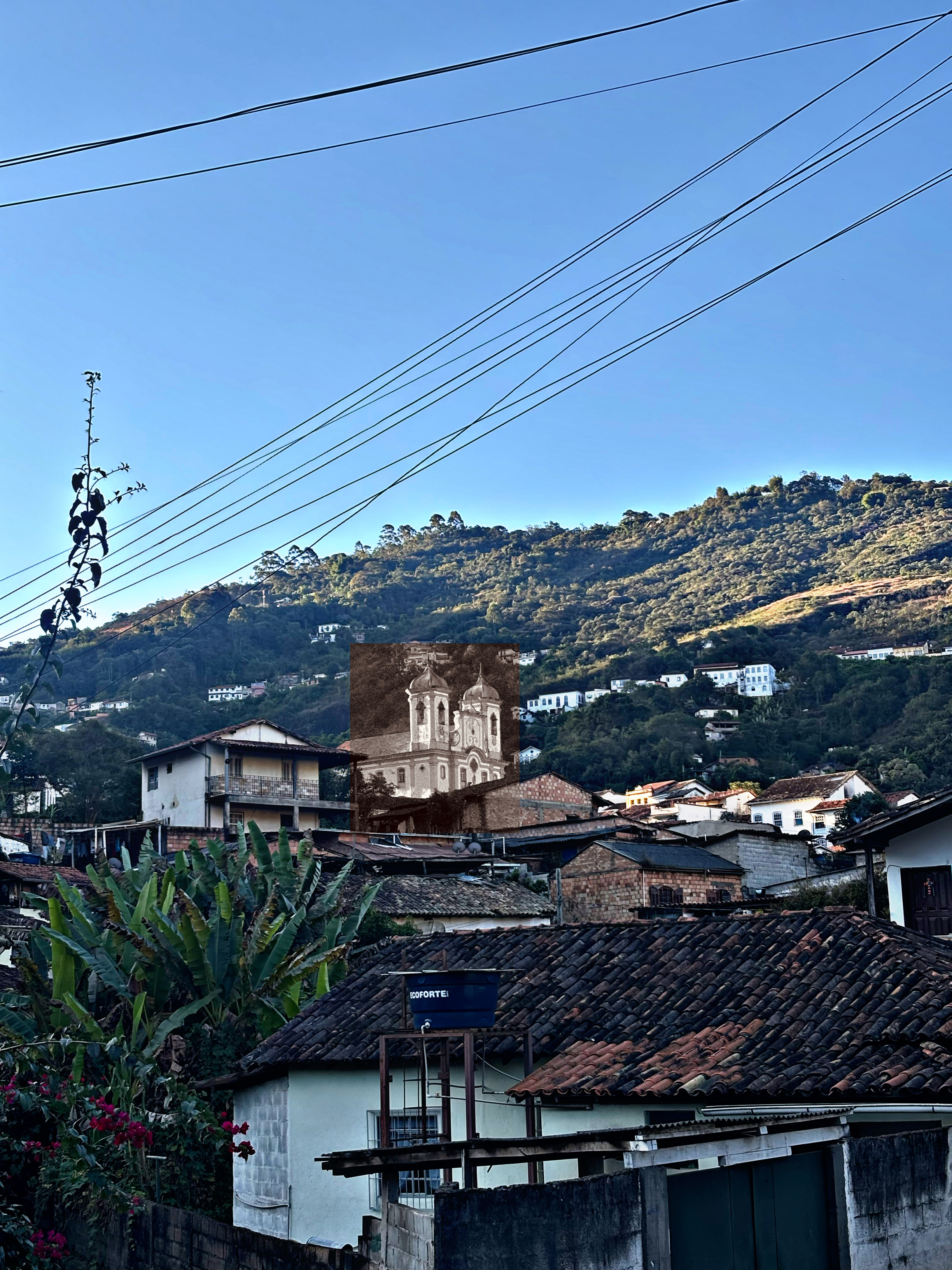 O Sol que ilumina e queima, de Pedro Lourenço Fotografia de uma paisagem urbana. Em primeiro plano, telhados de barro de casas simples, uma caixa d'água, folhagens e fios de eletricidade cruzando um céu azul claro. Ao fundo, uma montanha verde salpicada de casas. No centro da cena, destacando-se das construções, há uma inserção retangular em tom sépia mostrando uma igreja colonial de duas torres, contrastando com o restante colorido da foto.
