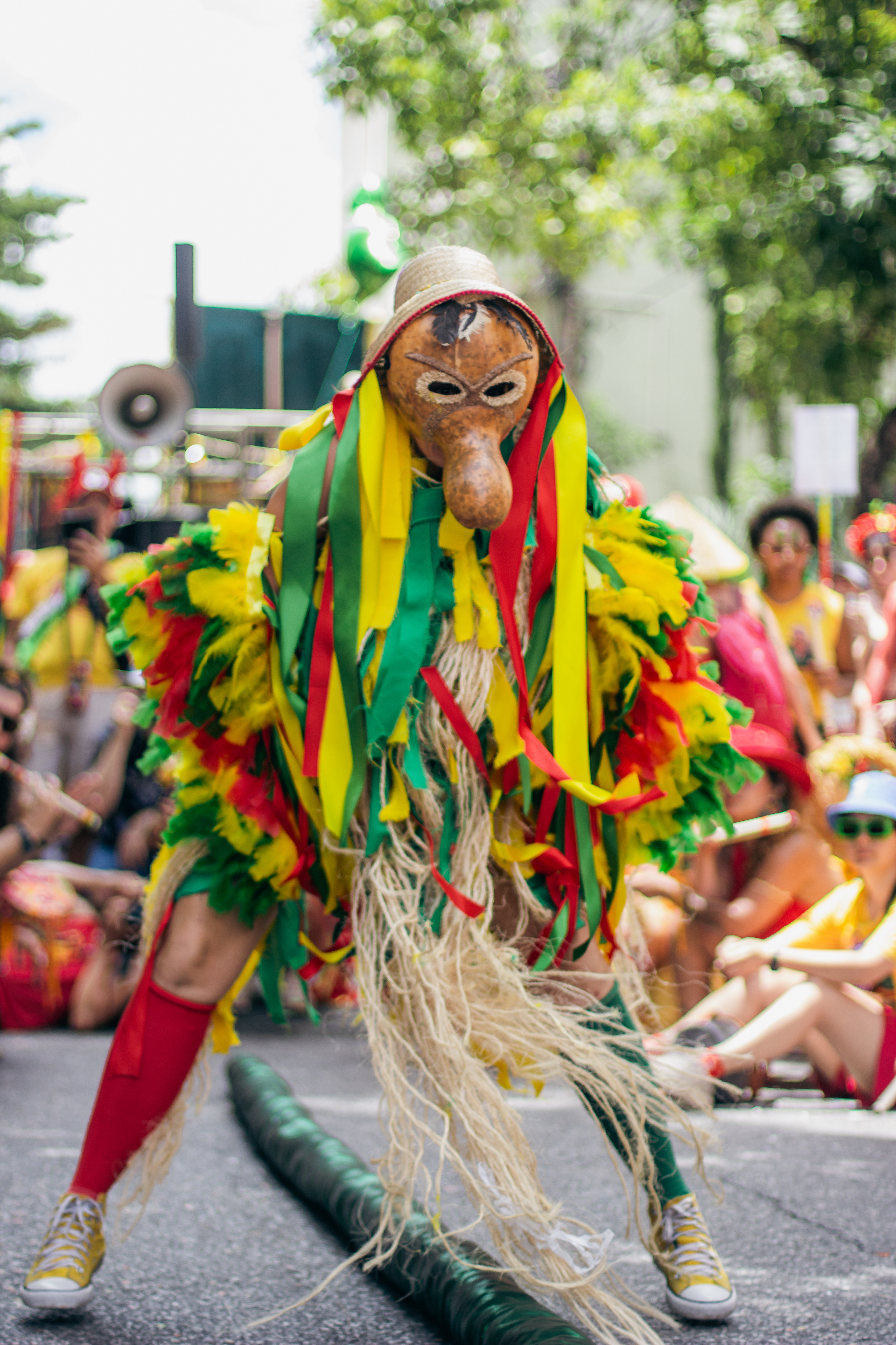 Fotografia colorida de um artista com traje performático agachado no centro de uma rua de asfalto. A pessoa usa uma máscara de cabaça rústica com nariz longo e um pequeno chapéu de palha. O traje é vibrante, coberto por franjas de tecido amarelas, verdes e vermelhas e fibras naturais longas. Usa tênis e meias altas coloridas. Ao fundo, uma multidão desfocada assiste sob árvores. No chão, um longo tubo verde.