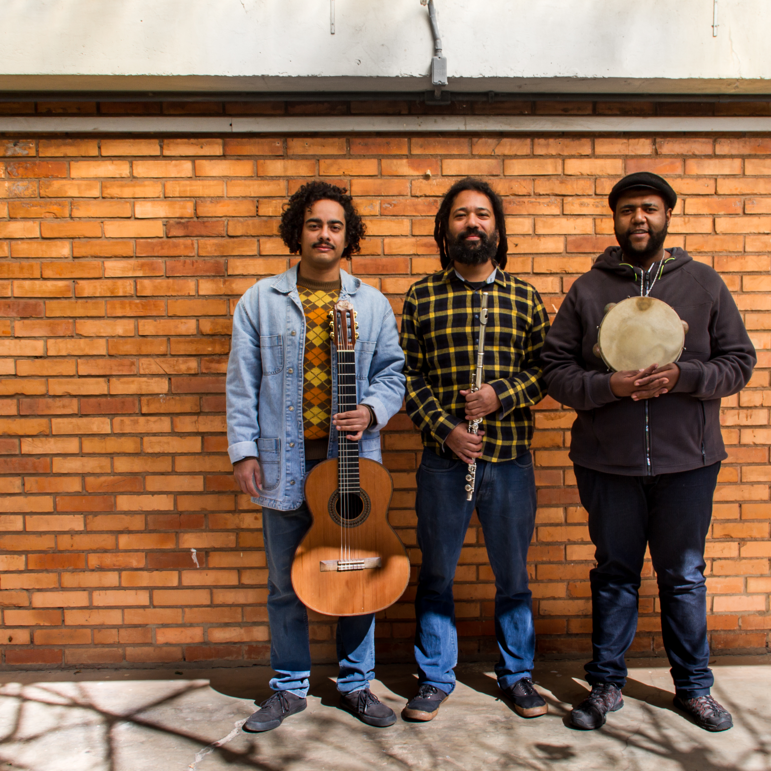 Fotografia do Trio Choro Negro Fotografia de três homens em pé, lado a lado, segurando instrumentos musicais em frente a uma parede de tijolos laranjas. O homem à esquerda veste jaqueta jeans e segura um violão. O do meio usa camisa xadrez amarela e preta e segura uma flauta transversal. O homem à direita veste casaco escuro, boina e segura um pandeiro com as duas mãos. O chão reflete sombras de galhos de árvores.